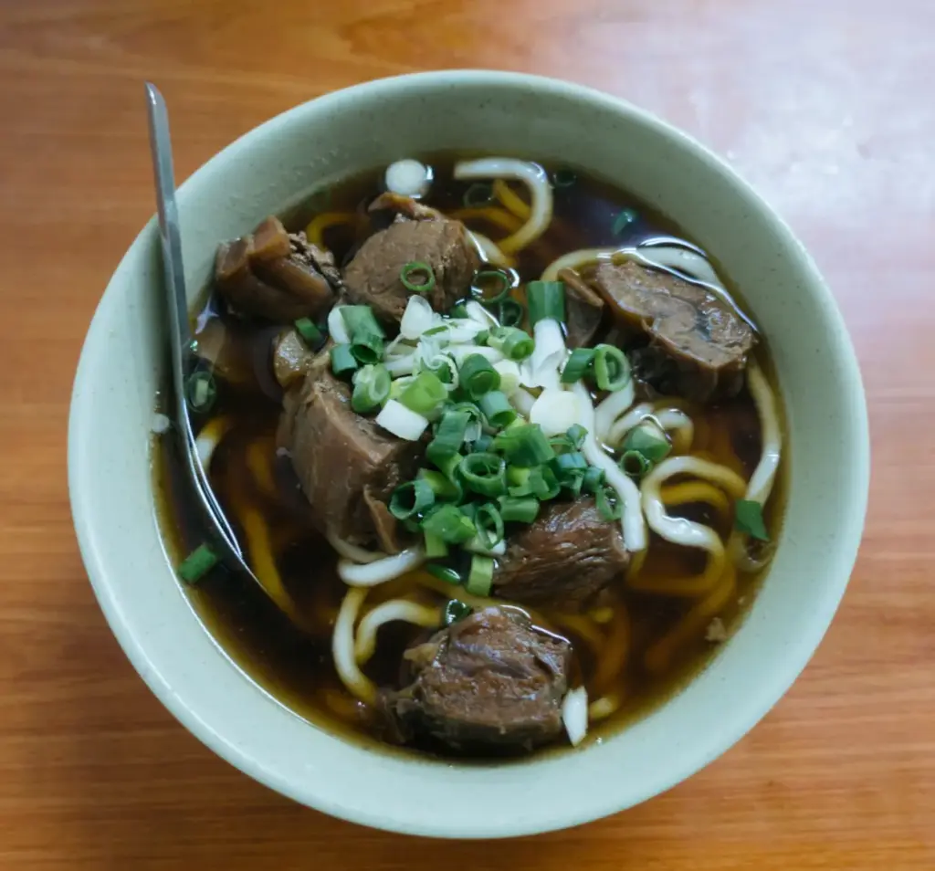 Taiwanese braised beef noodle soup with large tender beef chunks in a dark soy broth, thick wheat noodles, and sliced green onions