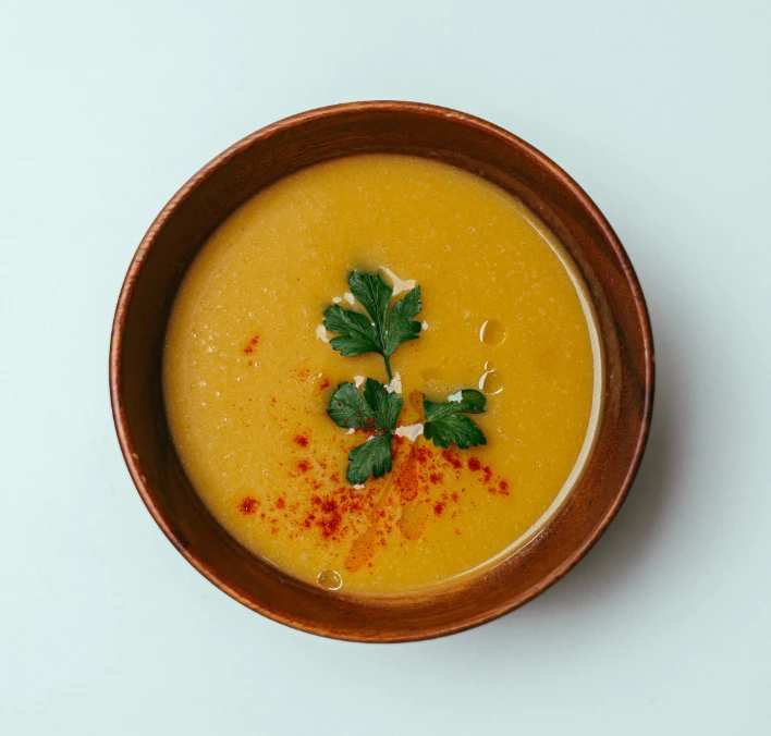 Bowl of golden creamy pumpkin soup in a rustic terracotta bowl garnished with fresh parsley and a dusting of red paprika on a pale blue background