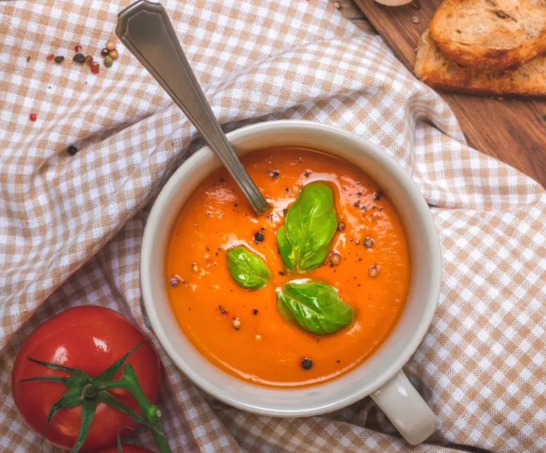 Bowl of vibrant orange-red Spanish gazpacho soup garnished with fresh basil and cracked pepper, with ripe vine tomatoes, a whole garlic bulb, and crusty bread on a rustic wooden surface