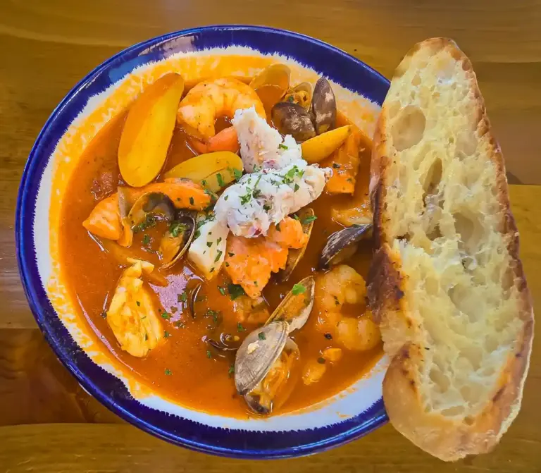 Spanish seafood soup Sopa de Mariscos with shrimp, clams, mussels, crab, and white fish in a saffron tomato broth, served in a blue ceramic bowl with toasted garlic bread