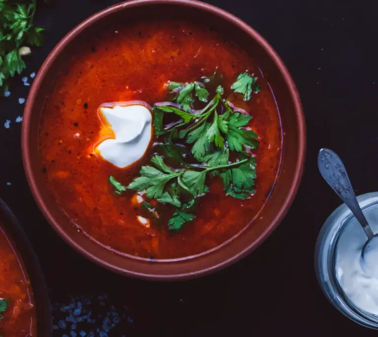 Ukrainian homemade borscht with deep crimson broth, topped with sour cream and fresh parsley, served on a dark surface with a jar of sour cream alongside